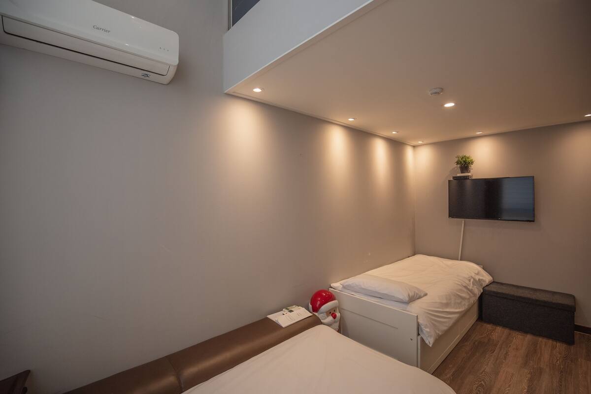 Una habitación de hotel moderna con una cama, un escritorio pequeño, un banco, un televisor montado en la pared y un equipo de aire acondicionado.