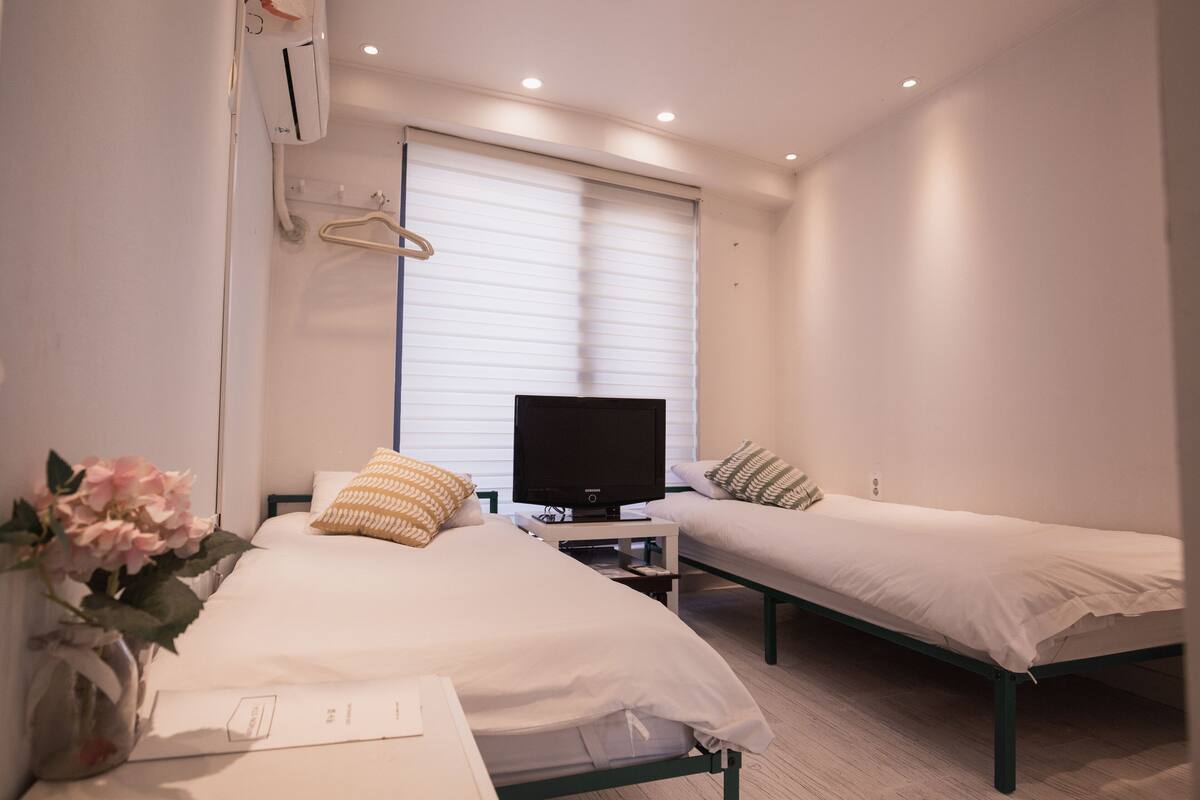 Habitación de hotel con dos camas individuales, un televisor sobre un soporte, un florero con flores y una ventana con persianas.