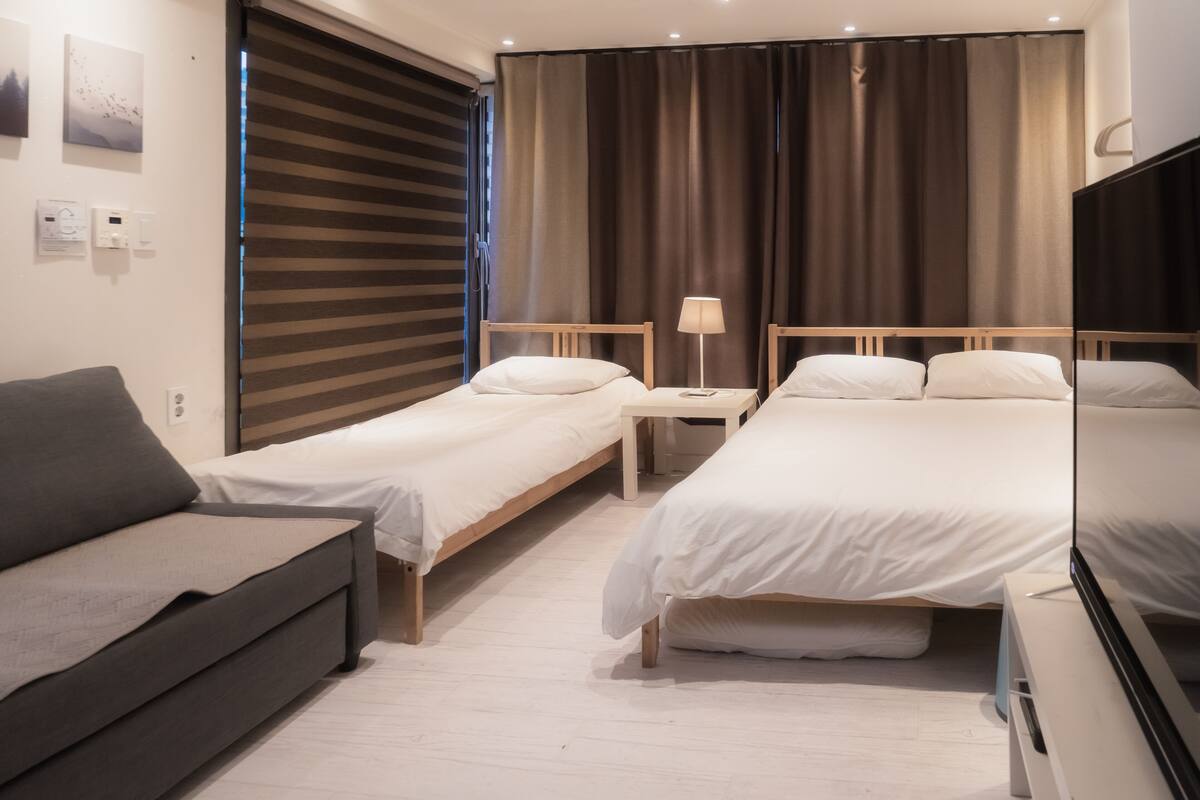 Habitación de hotel con dos camas individuales, un sofá, una mesita de noche con lámpara y un ventanal con cortinas.