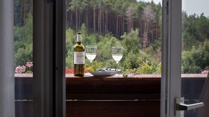 Una ampolla de vi, dues copes i un bol de fruita sobre una taula de fusta amb vista a un bosc a través d'una porta de vidre.