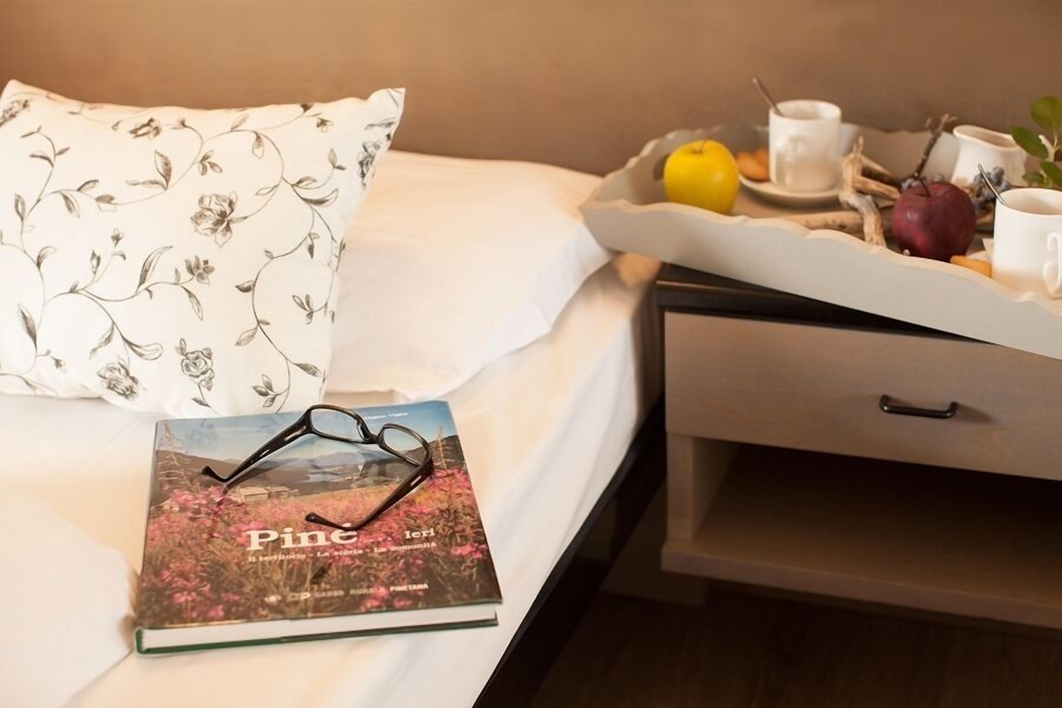 A neatly made bed with a floral pillow, a book titled 'Pine', a tray with fruits and a cup of coffee, and a drawer with a handle.