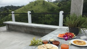 A table with food and drinks, a book, and a view of a green hill. - Hanthana House (Kandy)