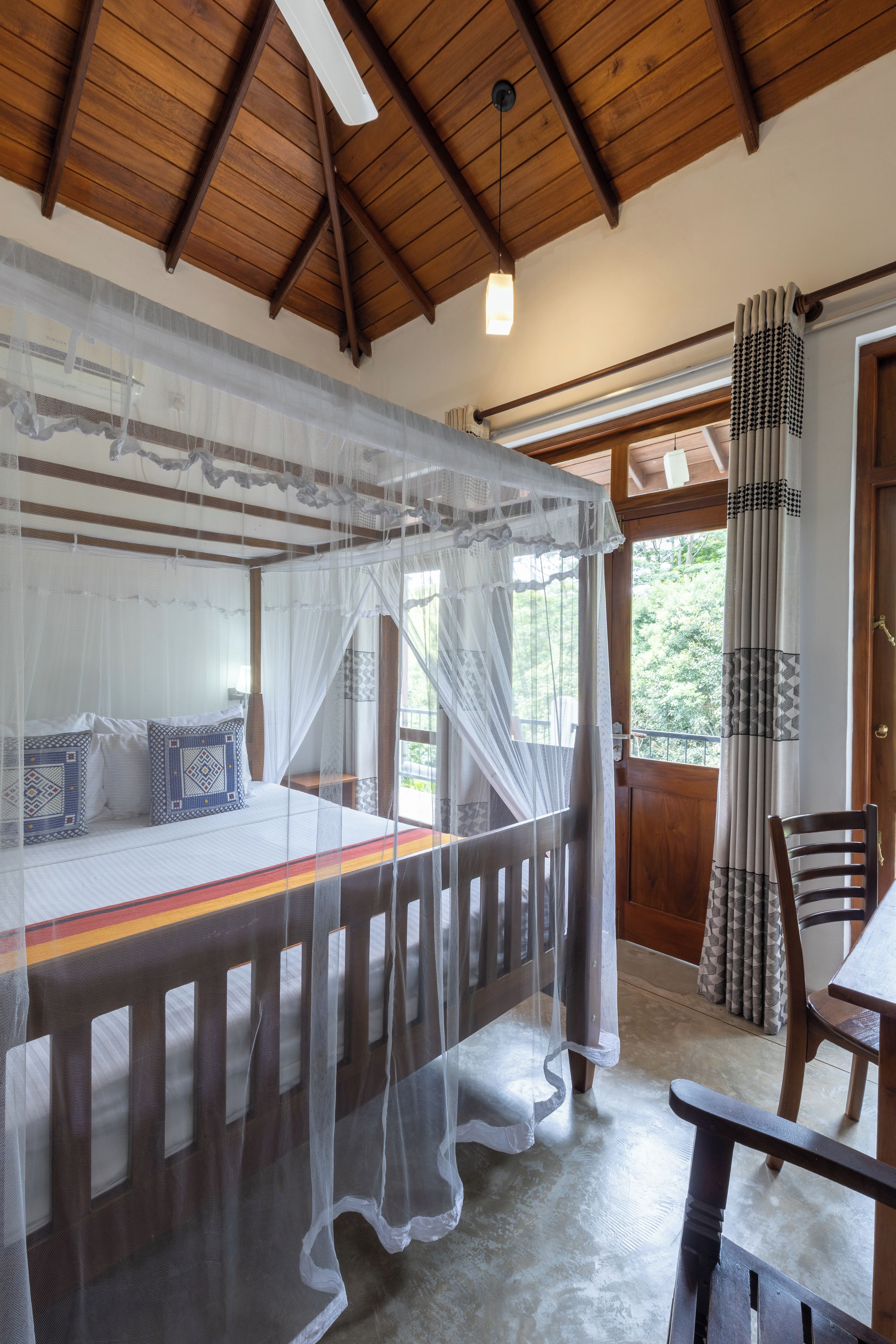 Una habitación con una cama bajo mosquitero, techo de madera, ventana con cortinas y puerta con cortinas estampadas.