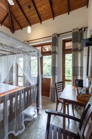 Quarto com teto de madeira, cama com mosquiteiro, mesa de jantar com cadeiras e vista para o verde lá fora.