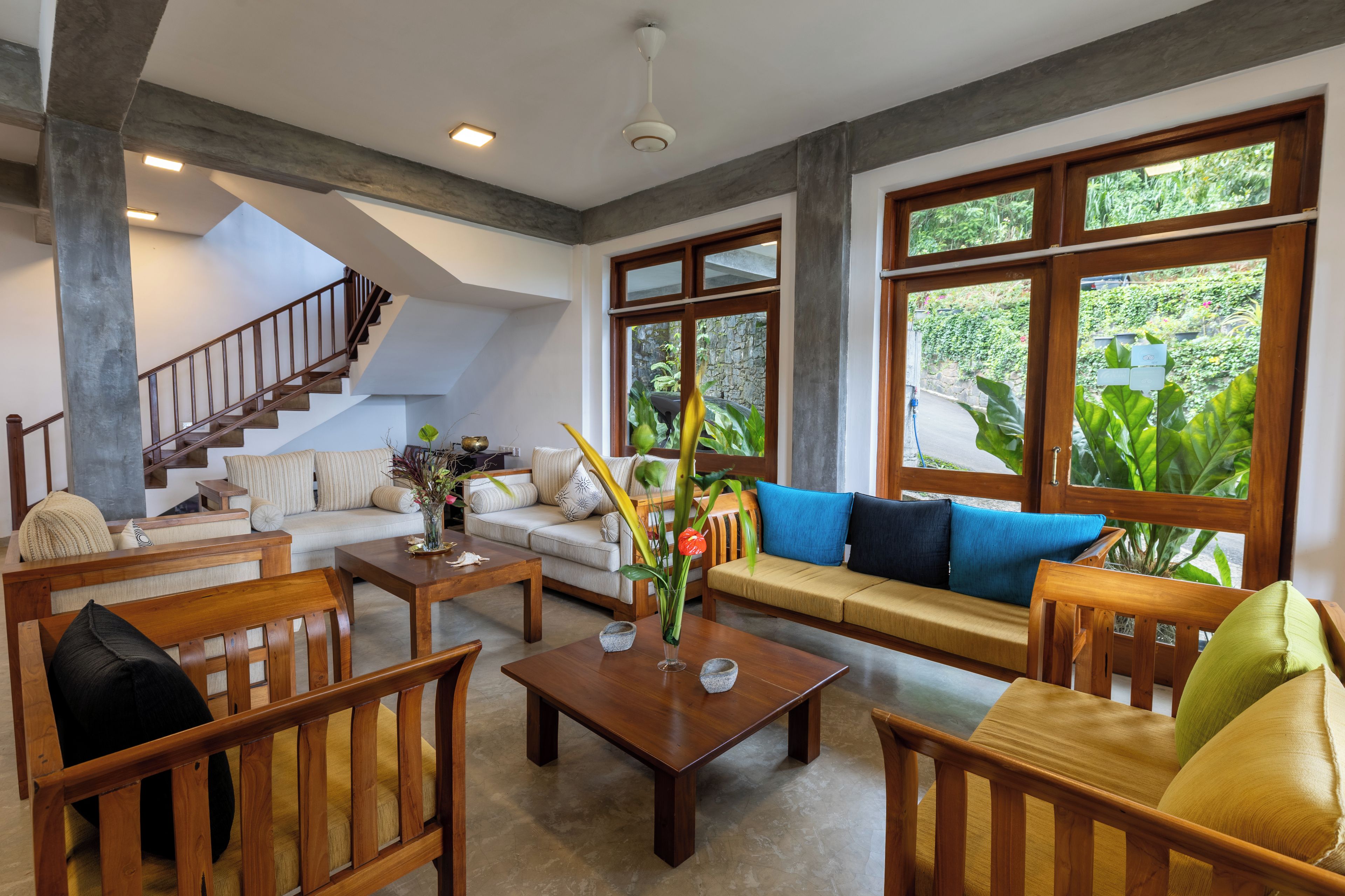 A spacious living area with a staircase, a sofa set, and large windows offering a view of greenery.