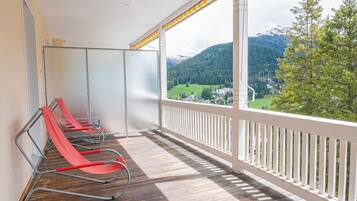 A balcony with a wooden floor and white railing, overlooking a mountainous landscape with a view of houses and greenery.