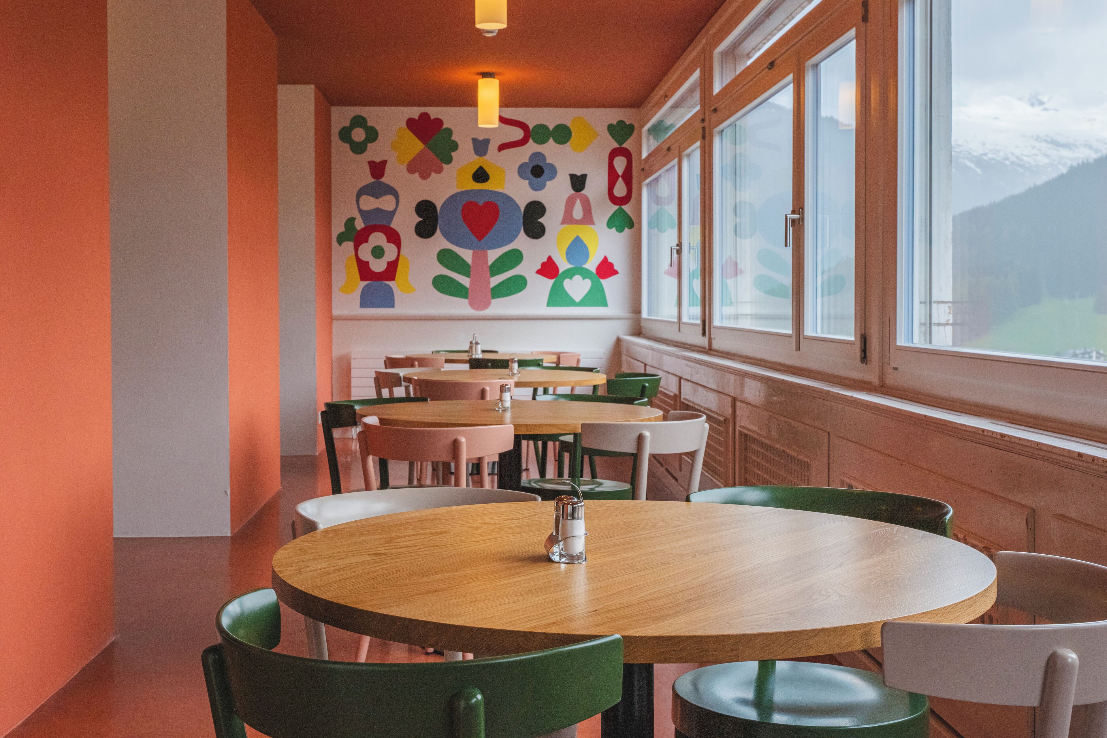 Un área de comedor con mesas de madera redondas y sillas verdes, un mural de pared colorido y grandes ventanales con vistas a un paisaje nevado.