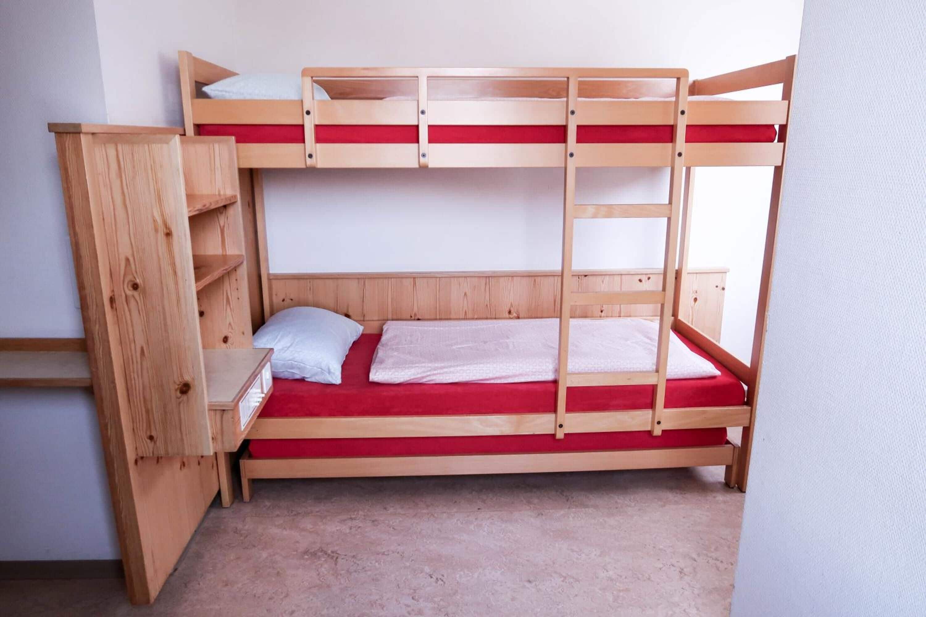 Una cama litera con escalera de madera, un estante pequeño y un colchón rojo.