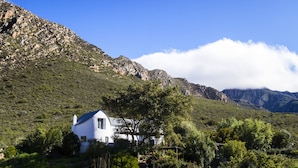 Une maison blanche avec un toit bleu, entourée de verdure et avec des montagnes en arrière-plan.