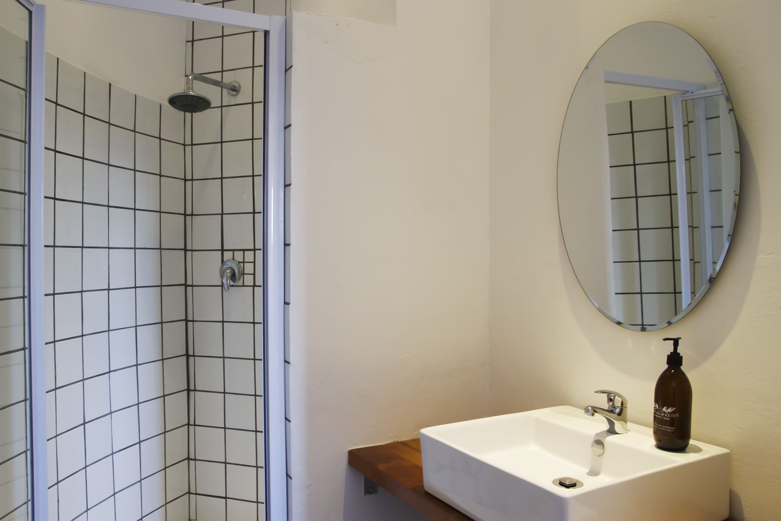 A bathroom with a white tiled shower, a white sink with a wooden shelf, and a round mirror.