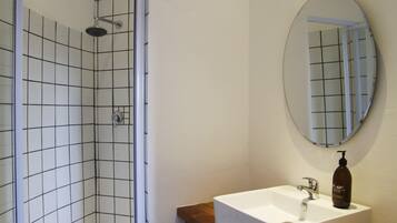 A bathroom with a white tiled shower, a white sink with a wooden shelf, and a round mirror.