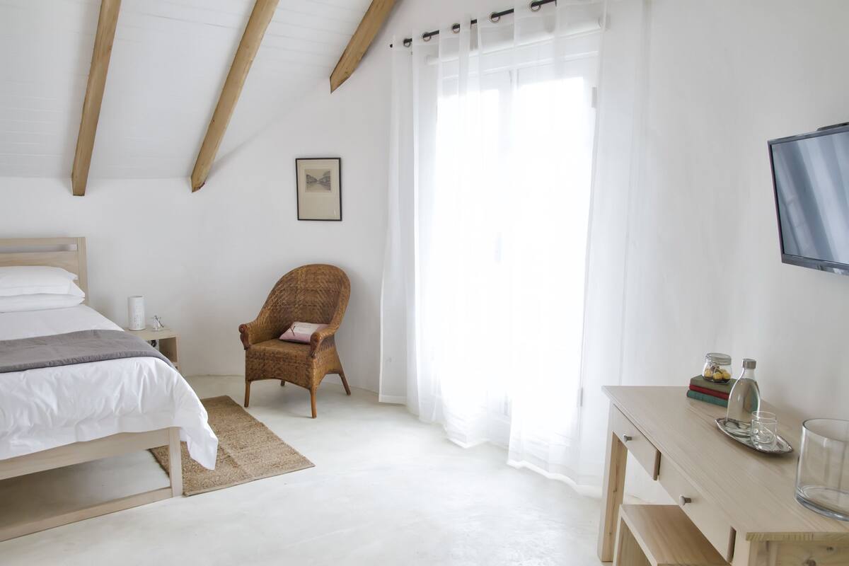 Ein Schlafzimmer mit Bett, Sessel, Schreibtisch, Fernseher und einem Fenster mit Vorhängen.