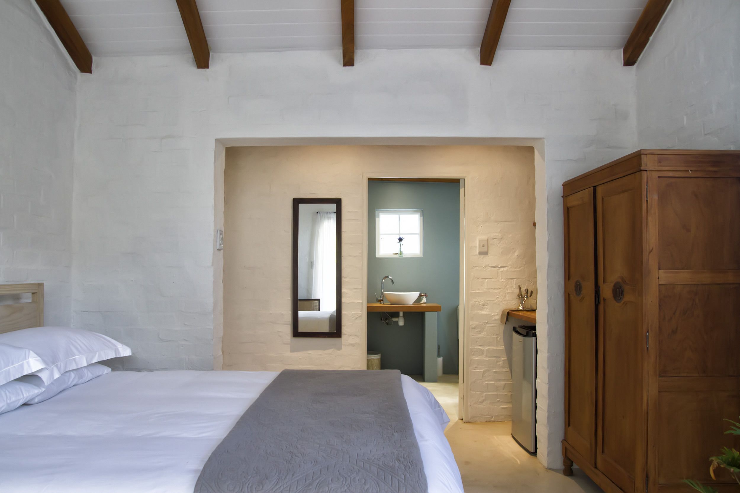 Une chambre à coucher comprenant un lit, une armoire en bois, une salle de bain avec un lavabo et un miroir, et une porte donnant sur une autre pièce.