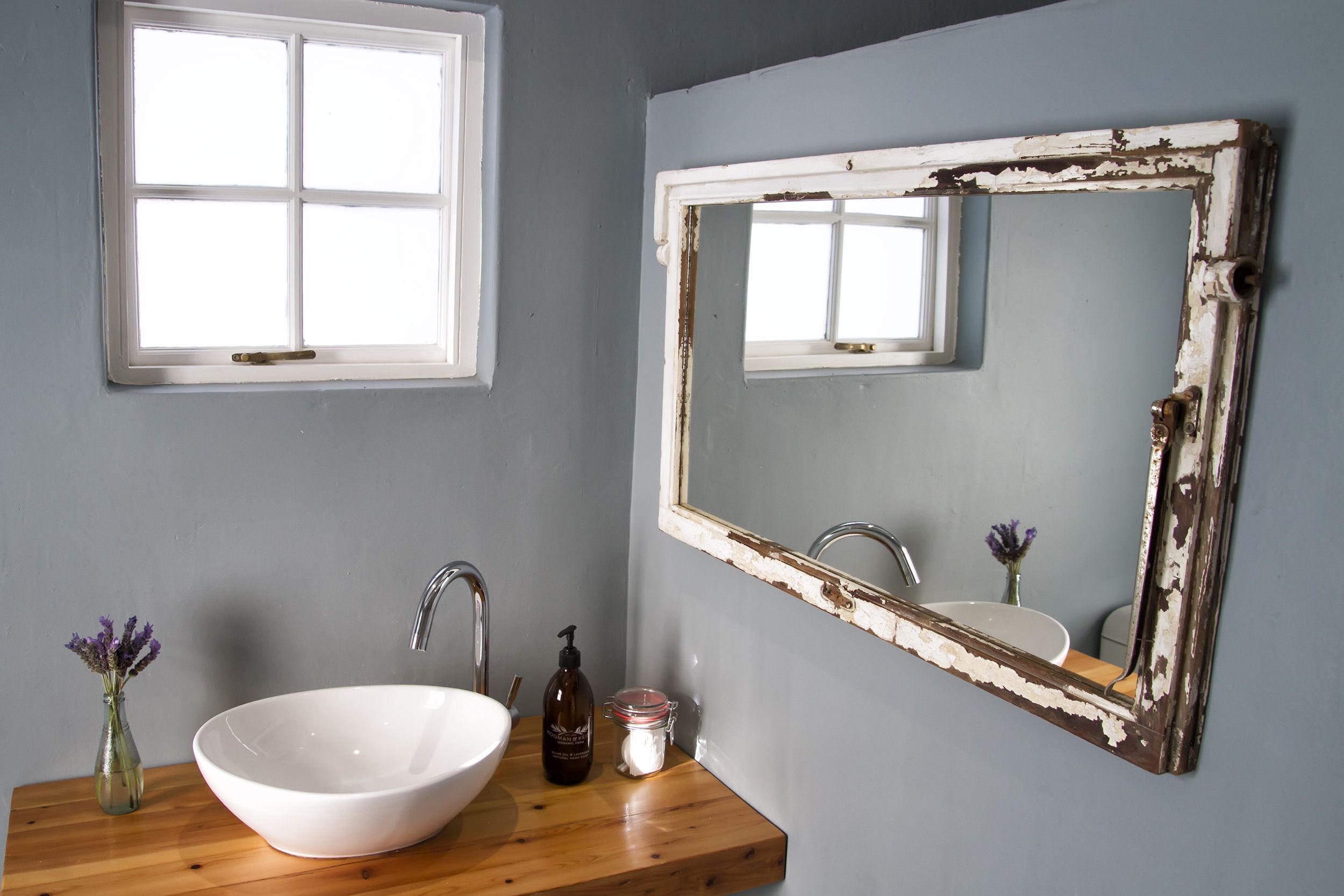 Une salle de bain avec un lavabo blanc, un plan de toilette en bois et un miroir dont la peinture s’écaille.