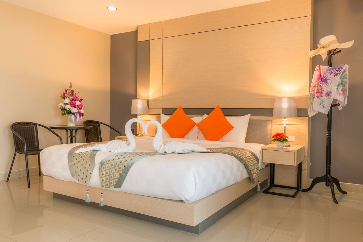 A neatly made bed with a swan-shaped pillow arrangement, a bedside table with a lamp, a chair, and a small table with a vase of flowers.