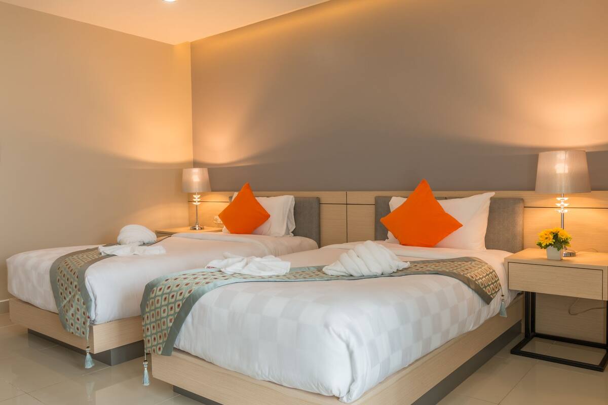 A double bed with white linens, orange pillows, and a bedside table with a lamp and flowers.