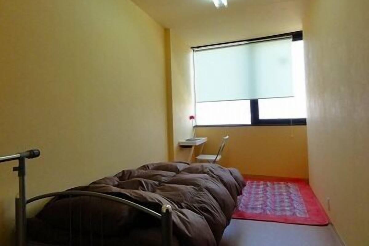 A small, narrow room with a single bed, a small table, and a window.