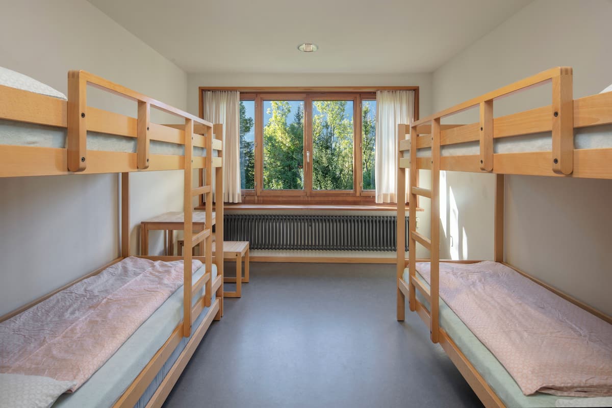 Ein Zimmer mit Etagenbetten, einem Vorhangfenster, einem Heizkörper und einem kleinen Tisch.