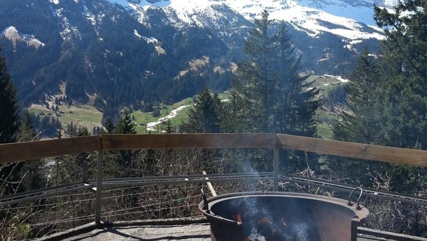 A stone patio with a metal fire pit in the foreground, overlooking a snow-covered mountain range and a valley with trees.