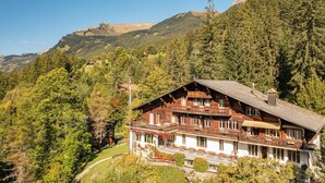 A large wooden chalet with multiple balconies, surrounded by dense forest and mountains.