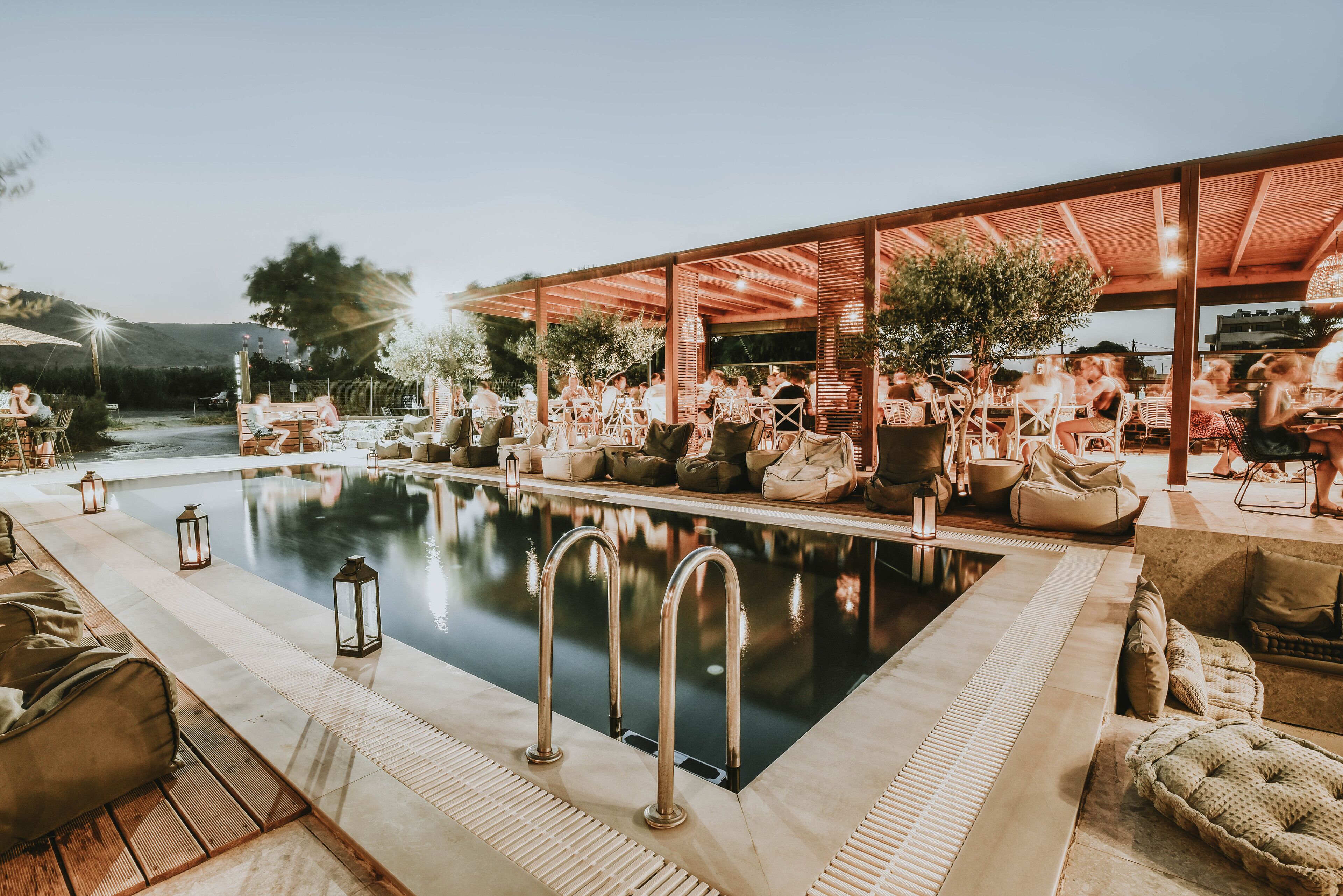 Zona junto a la piscina con sillones reclinables, mesas, una piscina y una zona de estar cubierta con árboles.