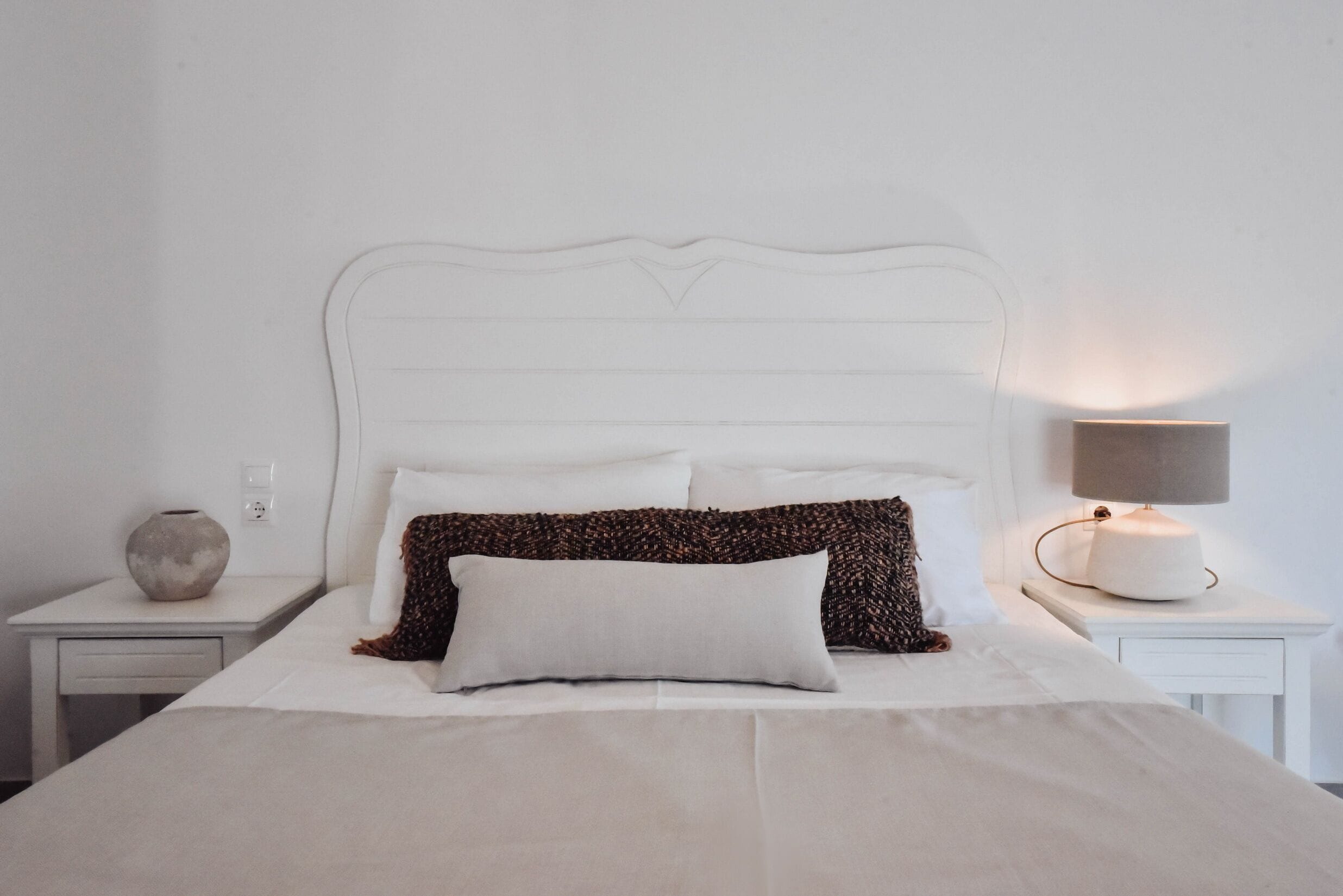 Una cama bien hecha con sábanas blancas, un cojín estampado y una lámpara de noche en una mesita blanca.
