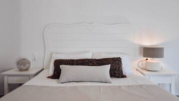 Una cama bien hecha con sábanas blancas, un cojín estampado y una lámpara de noche en una mesita blanca.