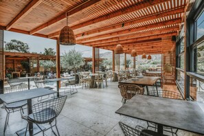 An outdoor cafe with wooden tables and chairs, a covered seating area, and large windows offering views of the surrounding landscape. - Ikaros Suites (Malevizi)