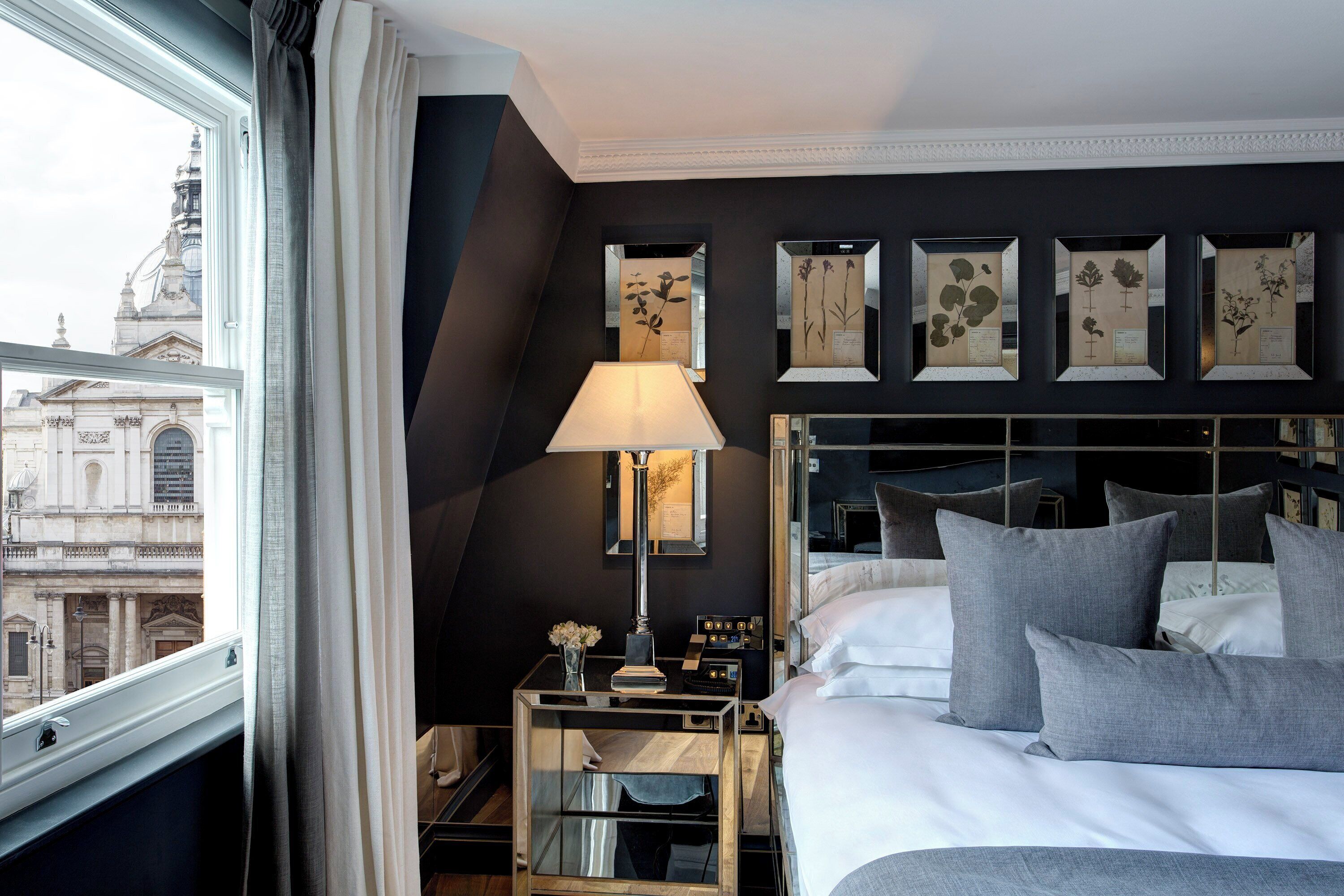 Habitación de hotel con cama, mesita de noche, lámpara e impresiones botánicas enmarcadas en la pared.