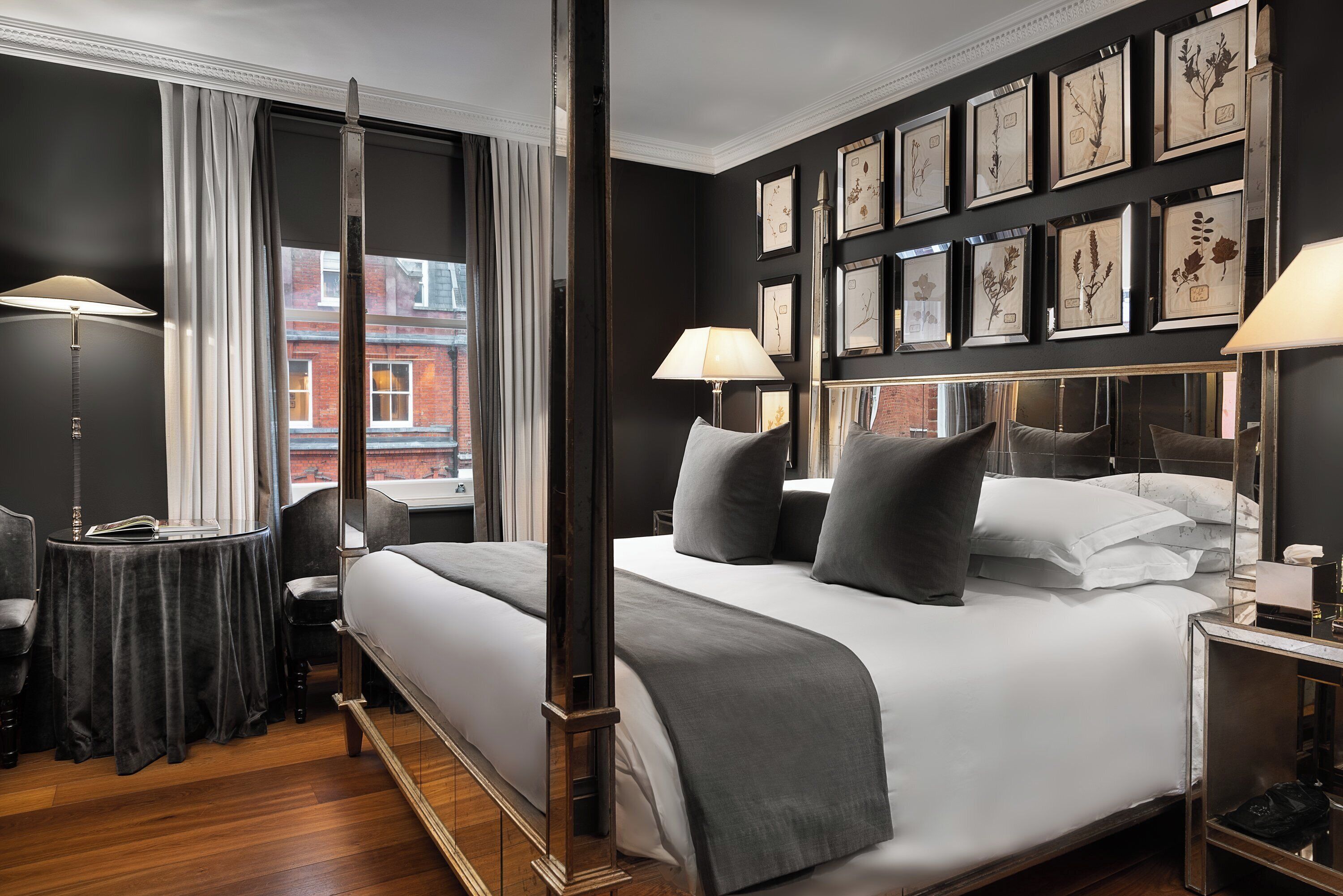 A four-poster bed with a grey and white bedding set, a bedside table with a lamp, and a wall adorned with framed botanical prints.