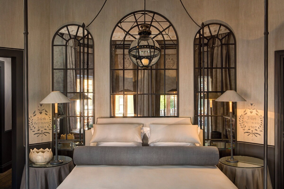 Habitación de hotel con una cama grande, dos mesitas de noche, un candelabro y ventanas amplias con vistas al exterior.