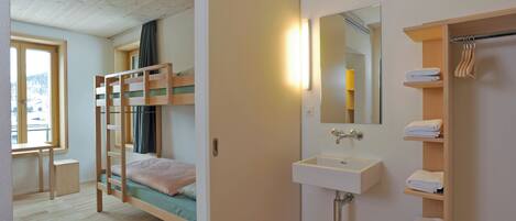 A bedroom with a bunk bed, a bedside table, a window, a bathroom with a sink and mirror, and a towel rack.