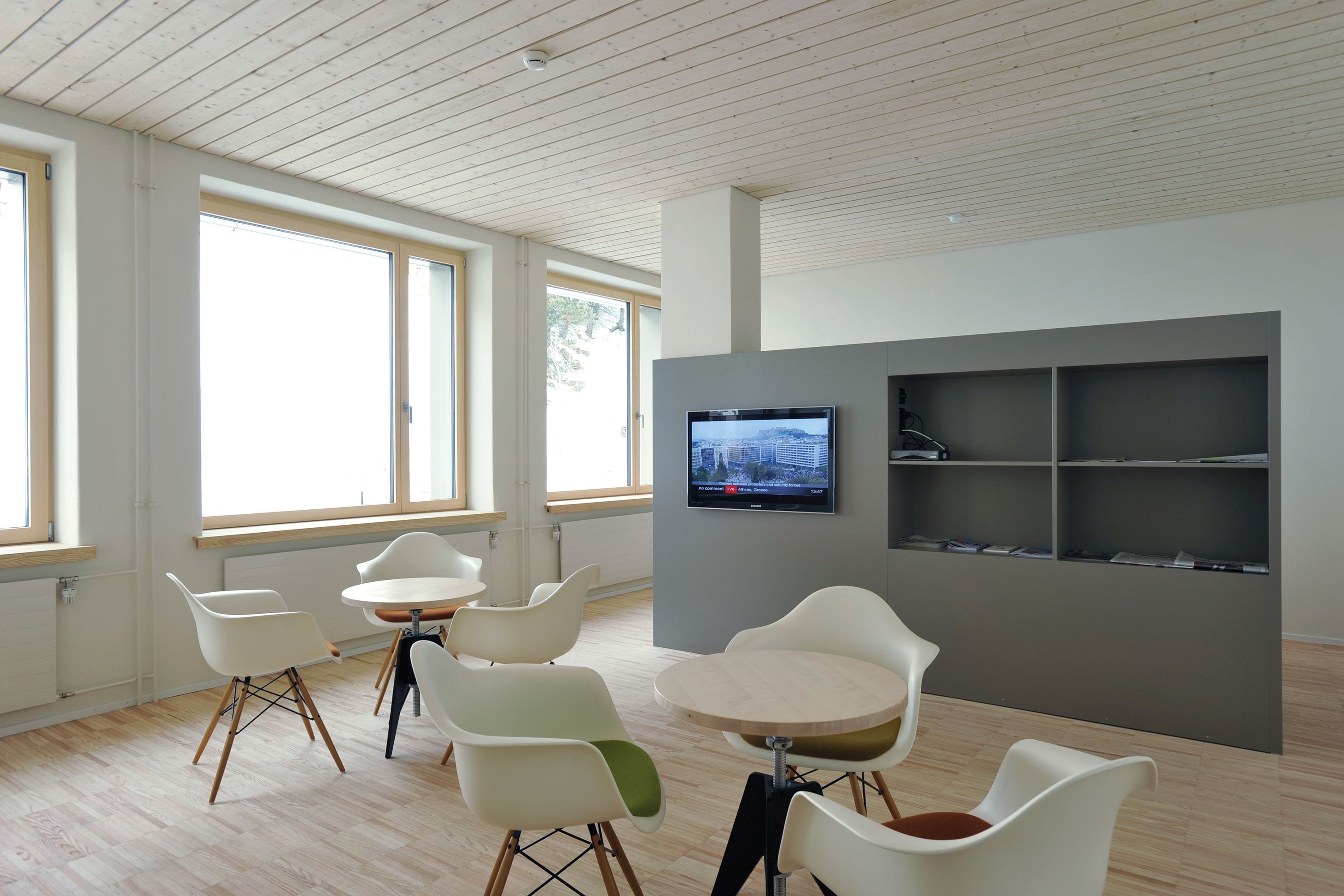 Una habitación moderna con ventanas amplias, sillas blancas, una mesa redonda y un televisor de pantalla plana montado en una pared gris.
