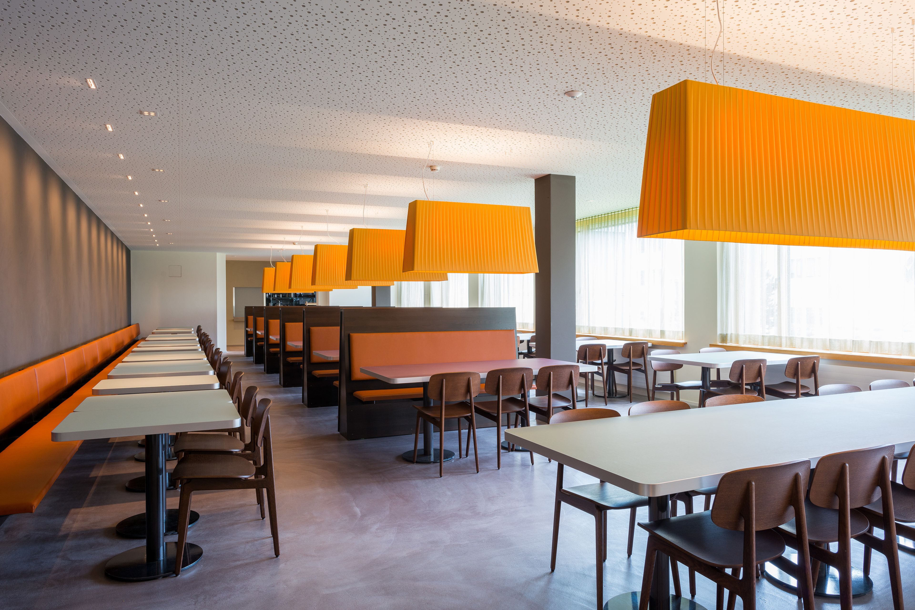 Un comedor moderno con mesas largas y cabinas, que presenta un techo moteado e iluminación colgante naranja.
