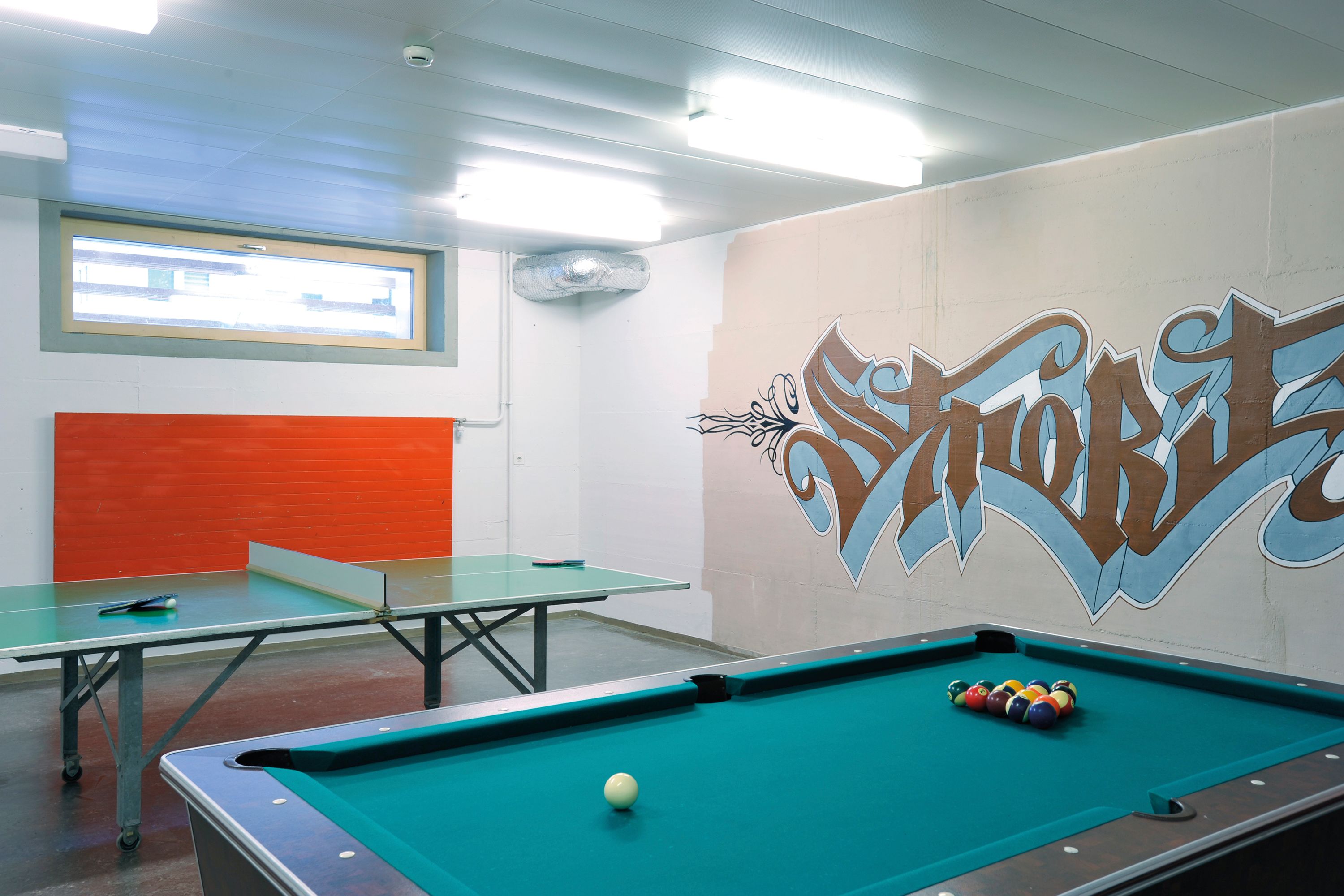 A billiards table with a pool cue and balls, a ping pong table, and a wall with graffiti art.