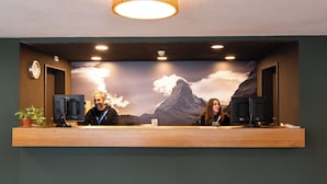 Un mostrador de recepción con dos monitores, un reloj y un gran mural en la pared con un paisaje montañoso.