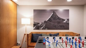 Una habitación con mesa de fútbol, un sofá con cojines, una lámpara y un cuadro de un paisaje montañoso en la pared.