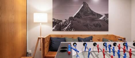A room with a foosball table, a sofa with cushions, a lamp, and a mountain landscape painting on the wall.