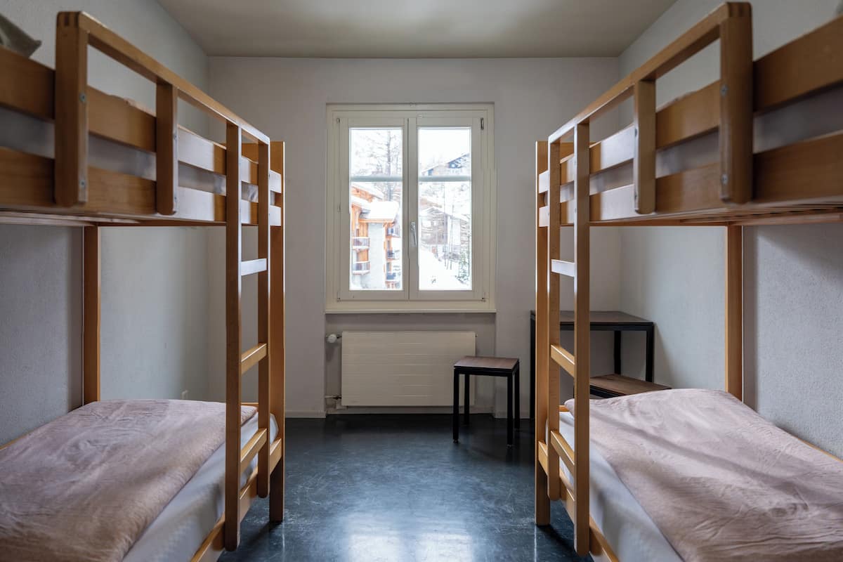 A dormitory room with bunk beds, a small table, and a window with a view of buildings.