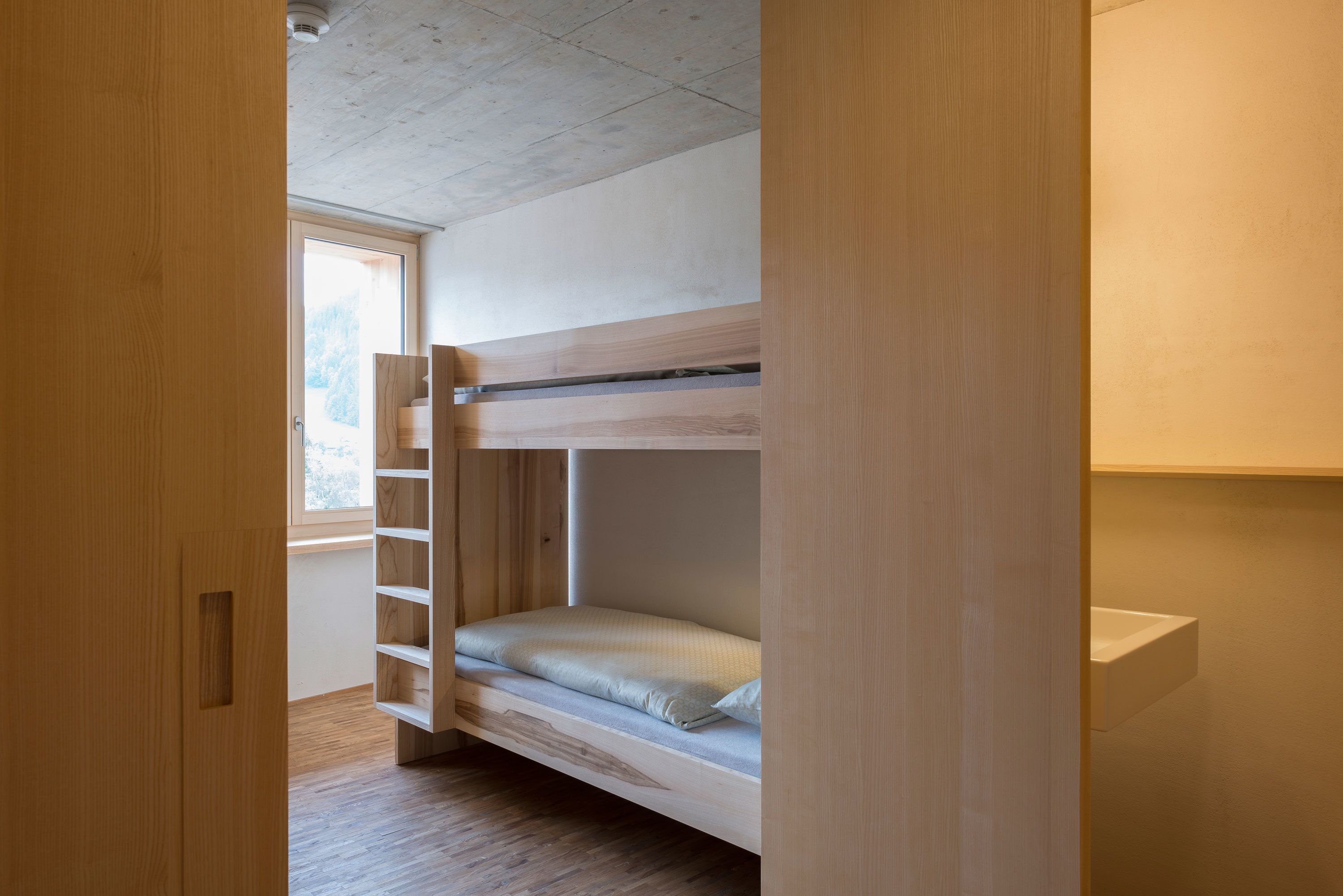 Una cama litera con escalera de madera, una ventana con vistas a árboles y un lavamanos blanco en una habitación pequeña.