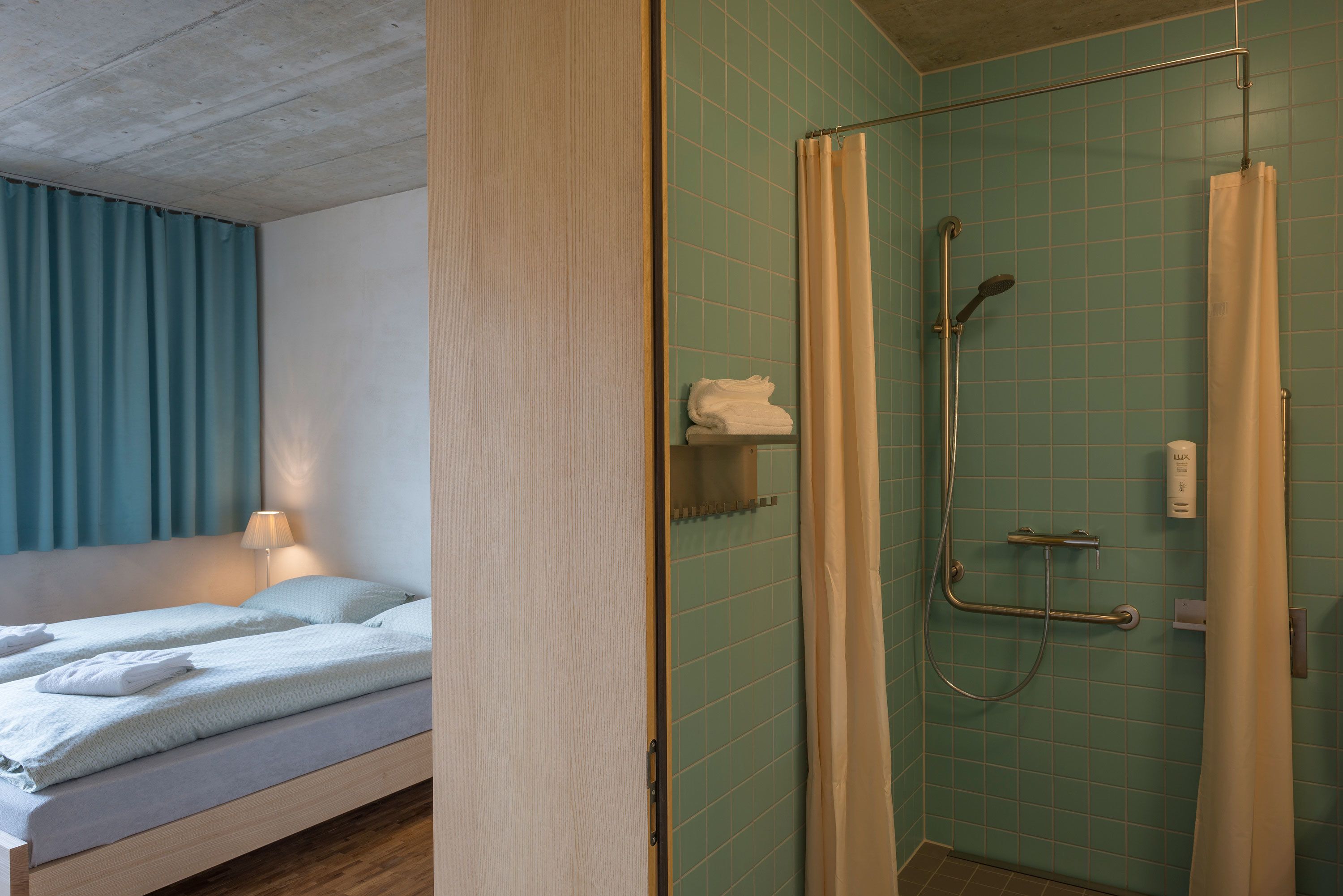 Quarto de hotel com cama, chuveiro com barra de apoio e porta-toalhas.