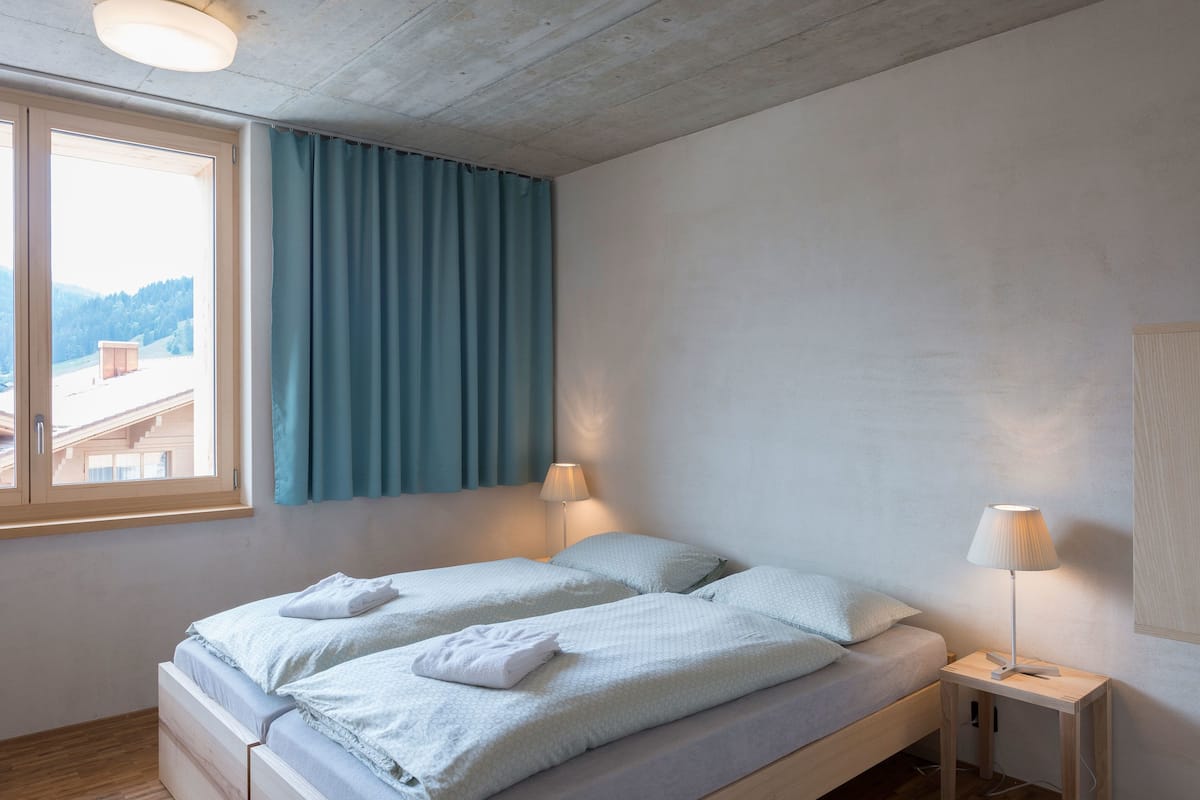 A bedroom with a bed, two bedside lamps, a window with curtains, and a view of a building and trees.