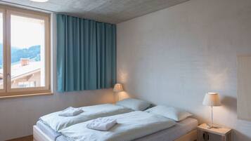 A bedroom with a bed, two bedside lamps, a window with curtains, and a view of a building and trees.