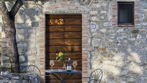 Una pared de piedra rústica con una puerta de madera, una ventana pequeña y una mesa con dos sillas y una botella de vino.