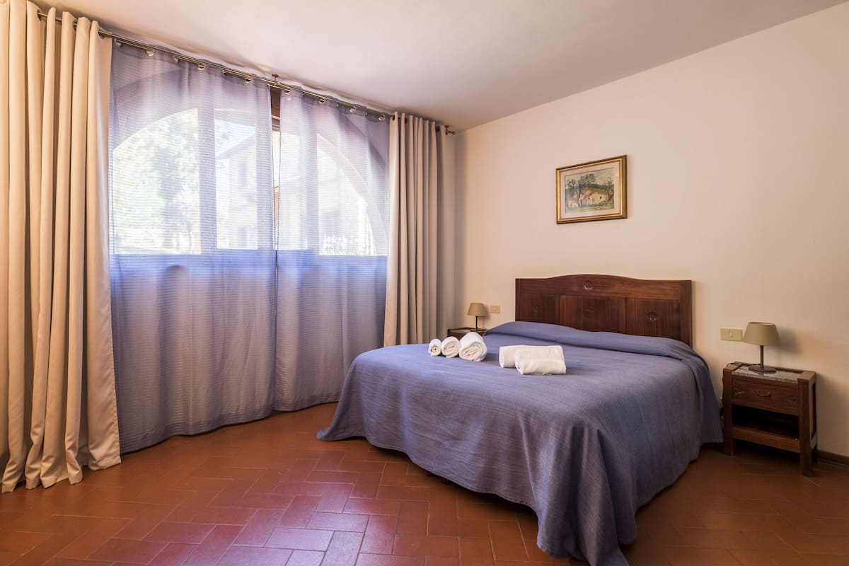 Une chambre à coucher avec un lit, des rideaux, une table de chevet, une lampe et un tableau au mur.