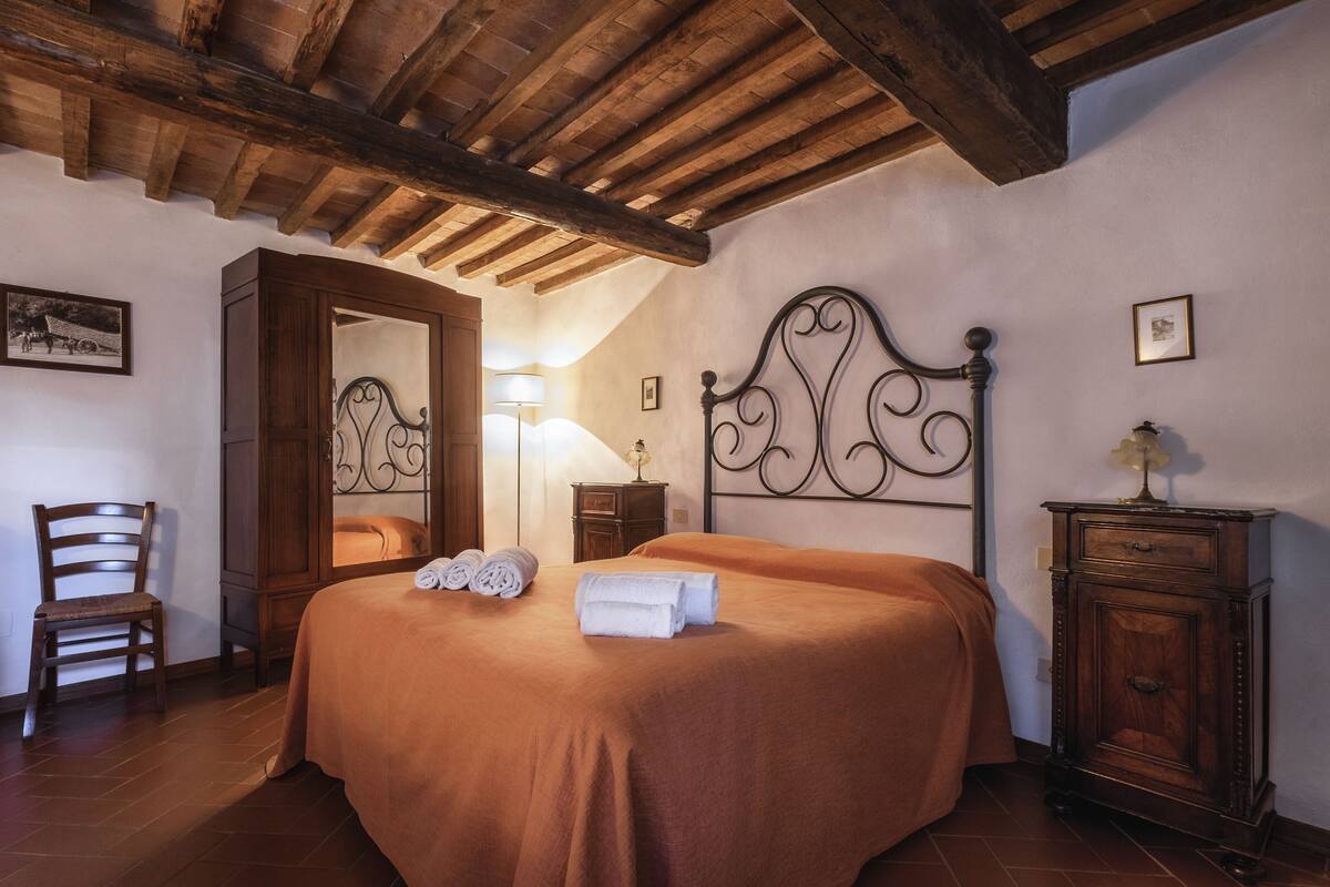 Une chambre à coucher avec un plafond en bois, un lit doté d’une tête de lit décorée, une table de chevet en bois, une chaise et une armoire.