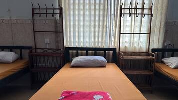 A dormitory room with two single beds, a central bed with a pink blanket, and a window with curtains.