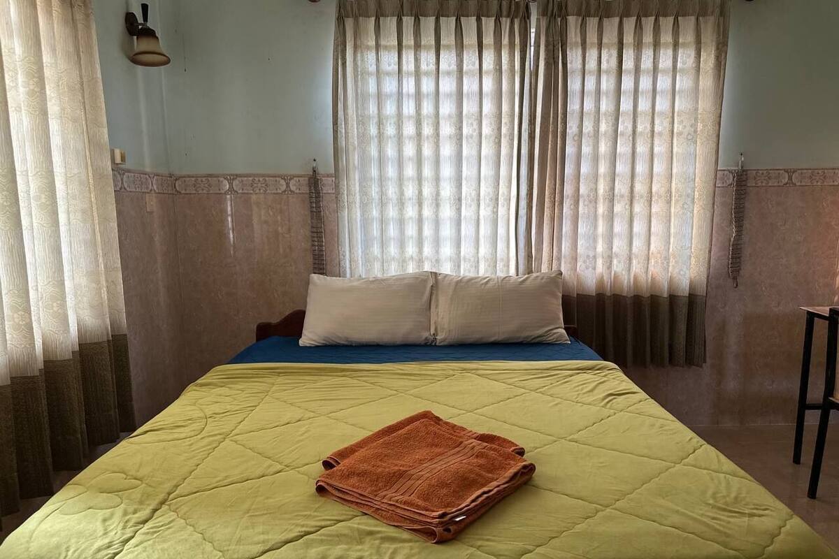 A neatly made bed with a green quilt, a folded brown towel, and two pillows on a blue mattress.