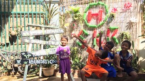 Un enfant, vêtu d’une tenue violette, se tient devant une clôture en bambou ornée de divers panneaux et décorations, dont un panneau indiquant « Local à linge ».