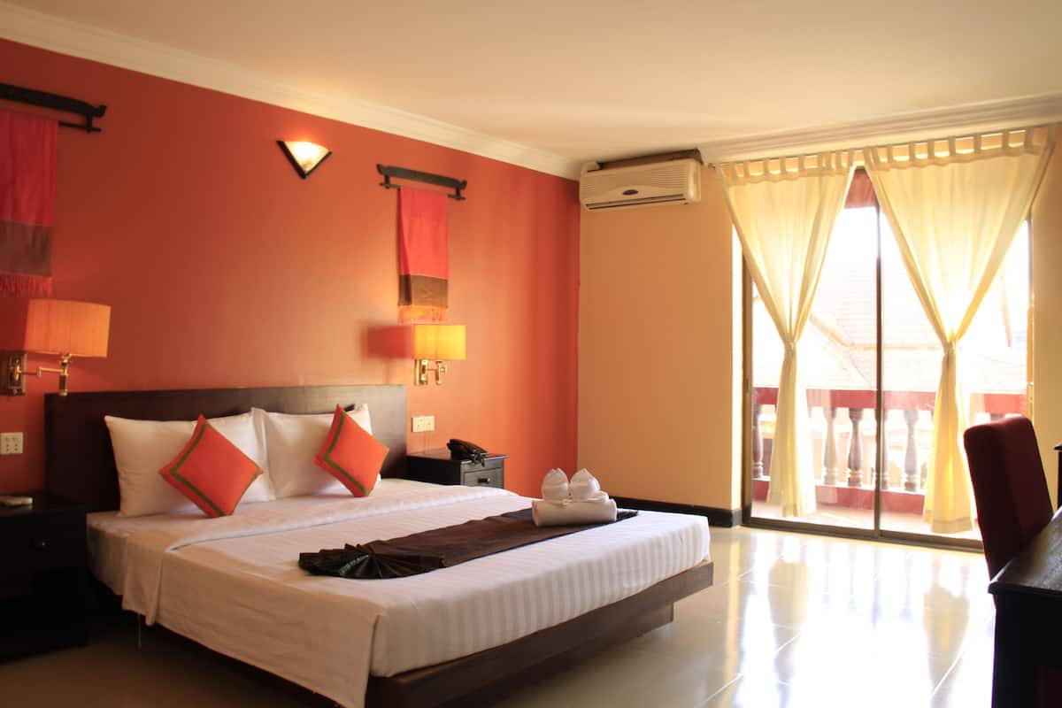 Une chambre d’hôtel avec un grand lit, une table de chevet, une lampe, un climatiseur mural et une vue sur l’extérieur grâce à une porte-fenêtre coulissante.